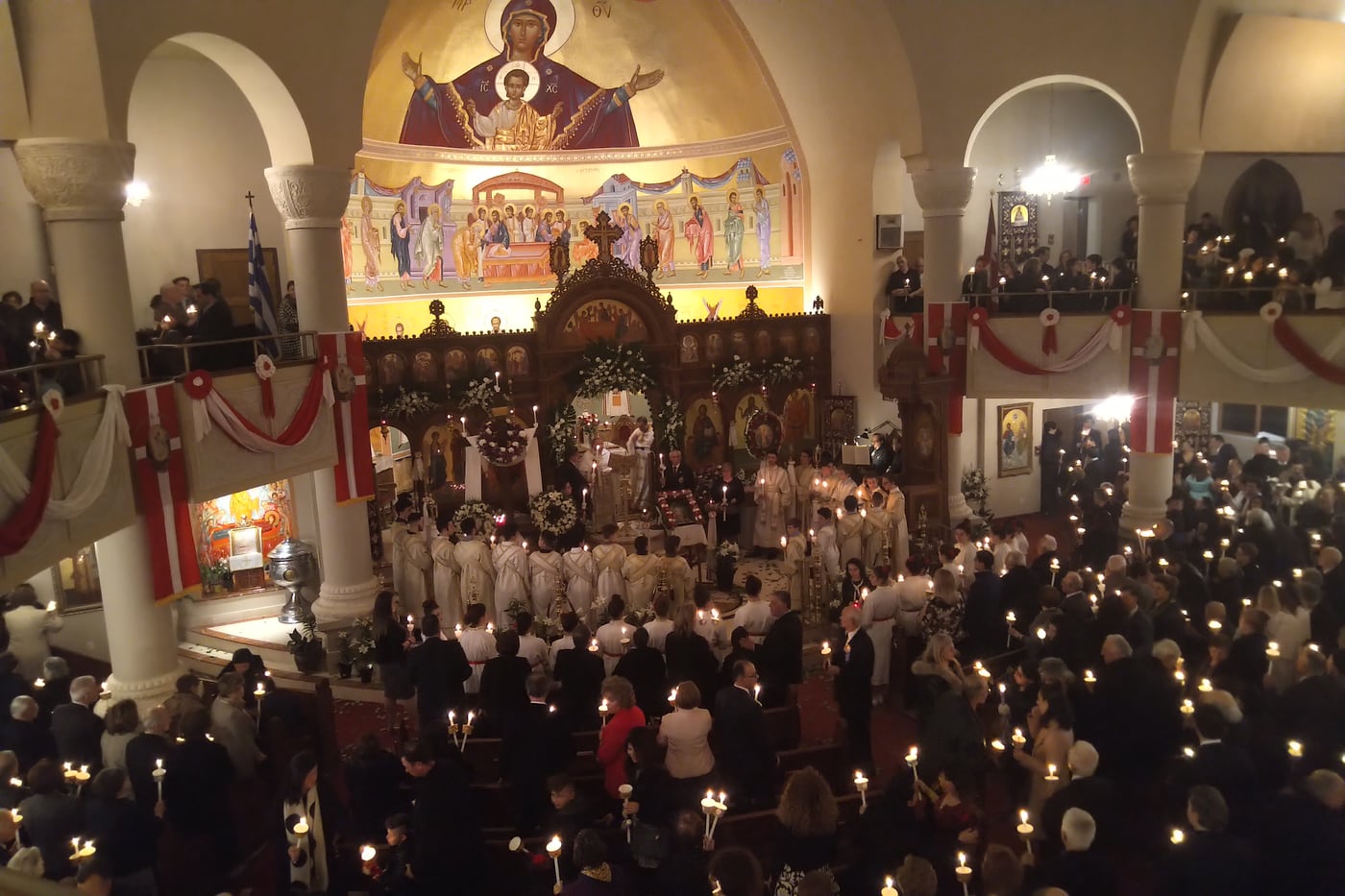 A church full of the faithful holding lit candles at the Orthodox Pascha service, the iconostasis dressed in white flowers.