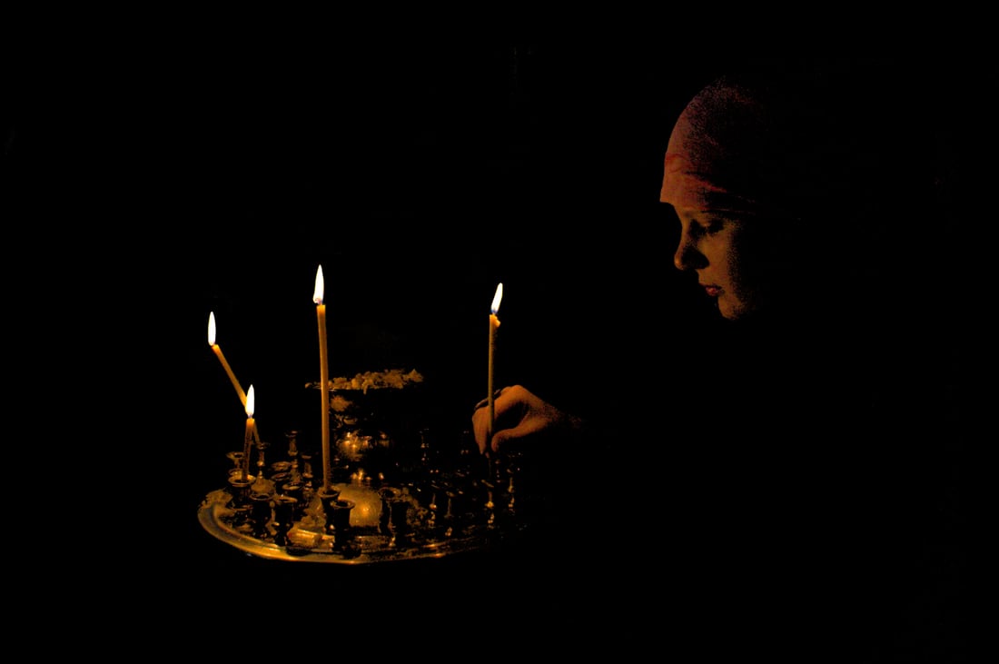 A hand lighting a thin beeswax candle before icons in the warm glow of an Orthodox church.